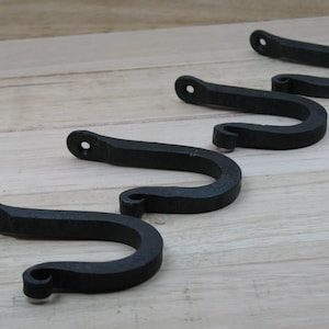 May include: Four black metal hooks with a decorative swirl at the end. The hooks are arranged in a row on a wooden surface.