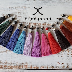 Può includere: Un set di 12 orecchini con nappe colorate, perline nere e dettagli argentati. Gli orecchini sono disposti in fila su una superficie di legno bianco. Il testo "Bunnyhood" è visibile al centro dell'immagine.
