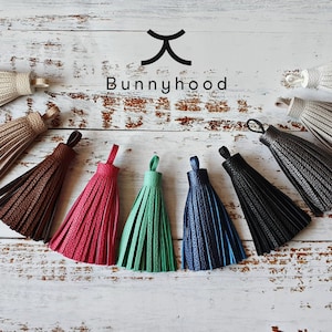 May include: Assortment of ten leather tassels in various colors, including white, beige, brown, pink, green, blue, and black. The tassels are arranged in a semi-circle on a white wooden surface. The text "Bunnyhood" is visible above the tassels.