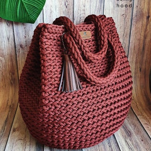 Può includere: Una grande borsa tote in crochet di colore rosso scuro con un fiocco in pelle marrone. La borsa ha un manico spesso a corda ed è realizzata con un motivo a punto testurizzato.