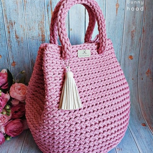 Puede incluir: Una bolsa de mano de crochet rosa con un borla de cuero blanco. La bolsa tiene un asa trenzada y una pequeña etiqueta de cuero con el texto "Bunnyhood" en ella.
