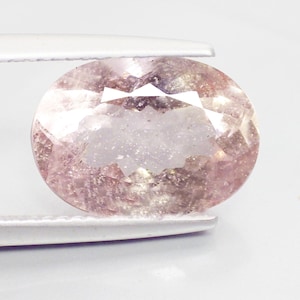 May include: An oval-cut gemstone held by tweezers. The gem has a pale pink hue with internal inclusions and facets that catch the light. The gemstone is set against a white background.