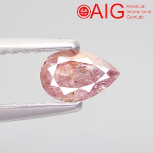 May include: A pear-shaped, light pink gemstone with visible internal inclusions and facets, held by silver-toned metal tweezers. The top right corner features the red AIG logo and the text American International GemLab in black lettering.