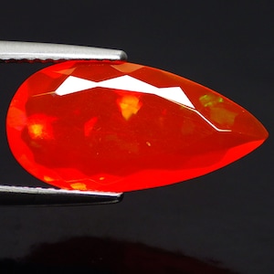 Puede incluir: Una gema facetada en forma de lágrima, sujeta con pinzas. La gema es de un color rojo anaranjado vibrante con reflejos de luz internos. El corte revela múltiples facetas, creando un efecto brillante. El fondo es negro.