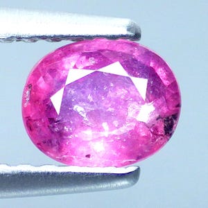 Puede incluir: Una gema rosa redonda y facetada sujeta por unas pinzas plateadas. La gema tiene un tono rosa vibrante con facetas rosas y blancas más claras. La gema es de talla brillante, reflejando la luz.