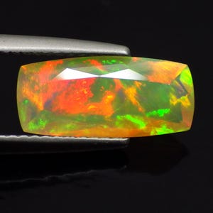 May include: A rectangular faceted opal gemstone displaying vibrant flashes of red, orange, and neon green play-of-color across its translucent surface. The stone features a polished finish with sharp geometric facets that catch the light.