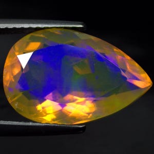 May include: A pear-shaped faceted opal gemstone displaying a vibrant play-of-color with intense flashes of electric blue, deep purple, and warm golden yellow hues throughout its translucent surface, held by dark metal tweezers.