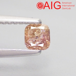 May include: A cushion-cut loose gemstone with a brownish-pink hue held by silver-toned metal tweezers. The stone features internal facets and light reflections. The logo for AIG American International GemLab is visible in the upper right corner.