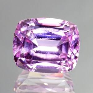 May include: A faceted, cushion-cut gemstone with a vibrant, light purple hue. The gem's facets reflect light, creating a sparkling effect. The gemstone is set against a reflective surface, enhancing its brilliance. This item is perfect for jewelry making.