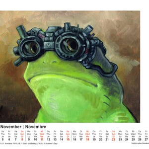 May include: A whimsical illustration of a green frog wearing steampunk goggles. The frog's face is centered, with the goggles prominently displayed. The background is a blend of brown and tan hues. A calendar for November is at the bottom.