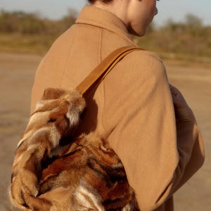 May include: A woman wearing a camel-colored coat carries a brown fur handbag with a leather strap.