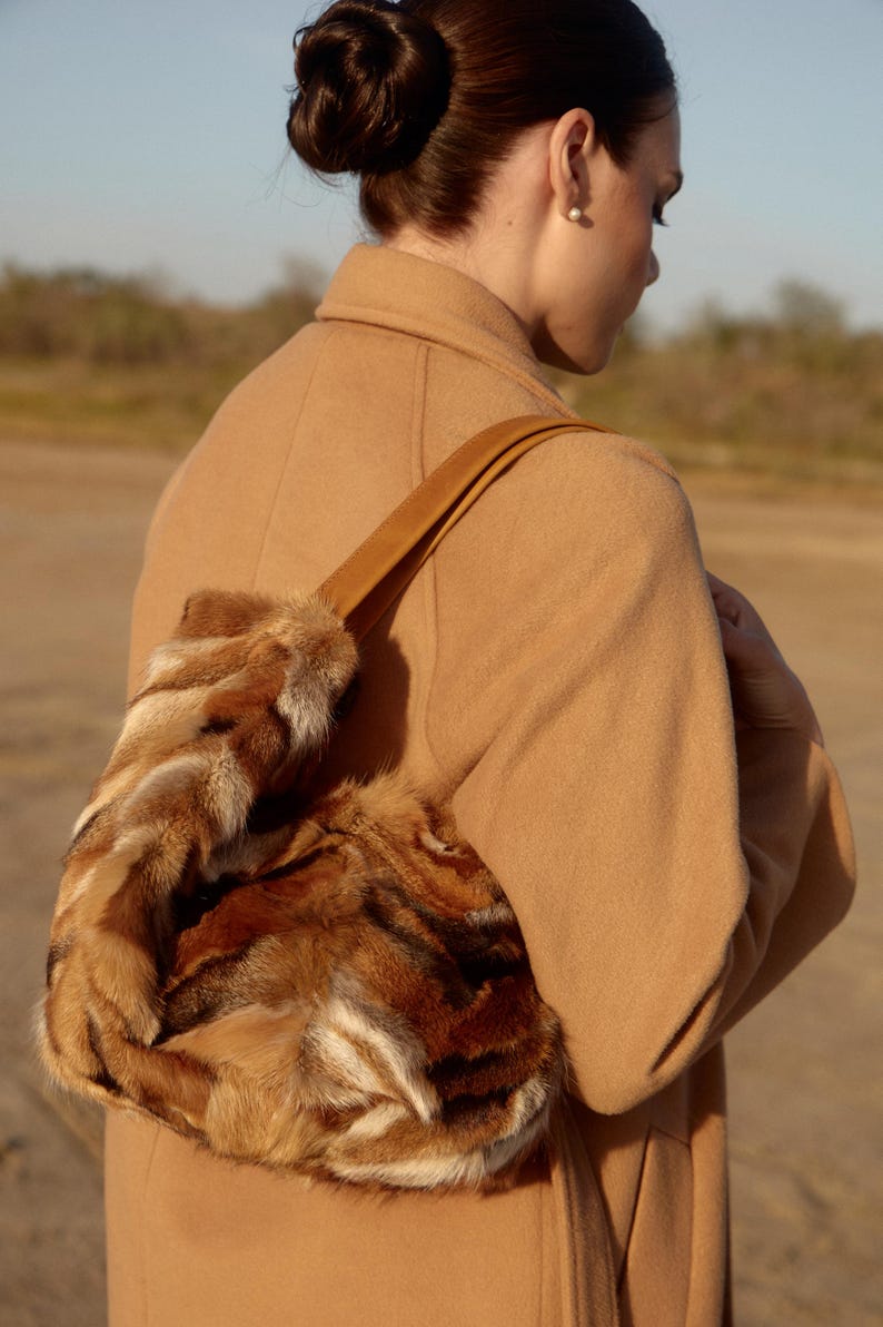 May include: A woman wearing a camel-colored coat carries a brown fur handbag with a leather strap.