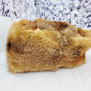 May include: A rectangular, golden-brown fur rug with a thick, textured pile. The rug is displayed on a white surface, with a snowy mountain backdrop. The fur has varying shades of brown and tan.