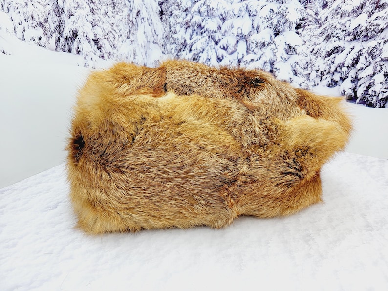 May include: A rectangular, golden-brown fur rug with a thick, textured pile. The rug is displayed on a white surface, with a snowy mountain backdrop. The fur has varying shades of brown and tan.