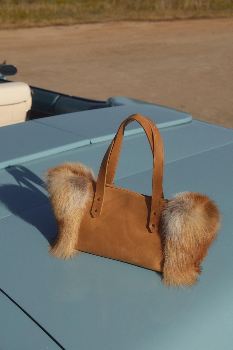 May include: A brown leather tote bag with fox fur trim. The bag is sitting on a blue car.