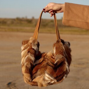 May include: A brown fur tote bag with leather straps. The bag is being held by a hand against a sandy background.