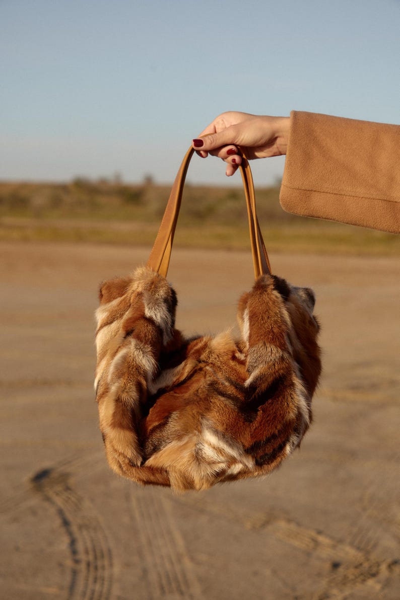 May include: A brown fur tote bag with leather straps. The bag is being held by a hand against a sandy background.