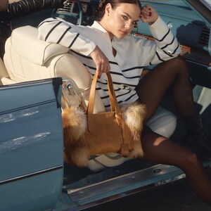May include: A woman wearing a white and blue striped sweater sits in a blue convertible car. She is holding a brown leather tote bag with a fur trim. The car is parked on a dirt road.