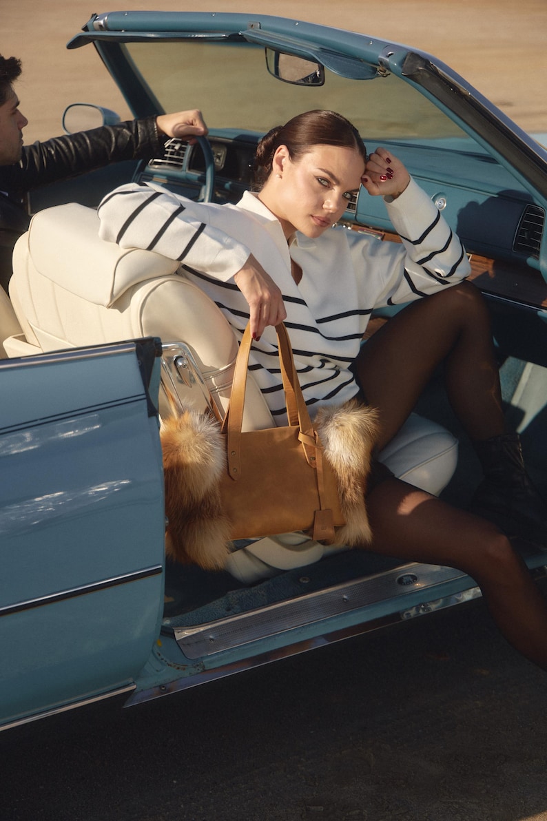 May include: A woman wearing a white and blue striped sweater sits in a blue convertible car. She is holding a brown leather tote bag with a fur trim. The car is parked on a dirt road.