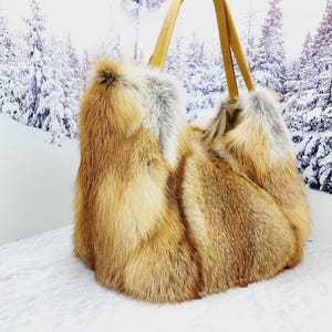 May include: A large, fluffy tote bag made of reddish-brown and gray fur. The bag has two tan leather handles. The background features a snowy winter scene with evergreen trees.