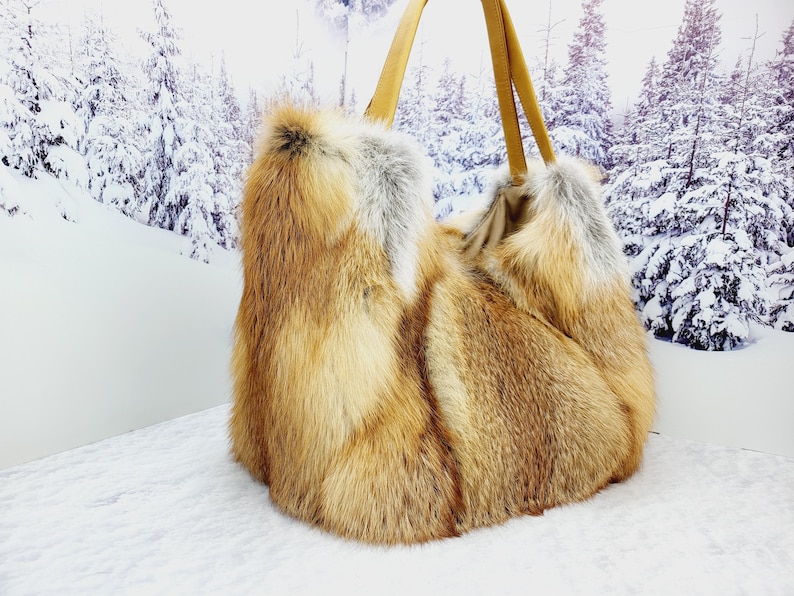 May include: A large, fluffy tote bag made of reddish-brown and gray fur. The bag has two tan leather handles. The background features a snowy winter scene with evergreen trees.