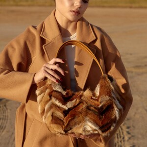 May include: A woman wearing a camel-colored coat holds a brown and white fur handbag with a brown leather strap.