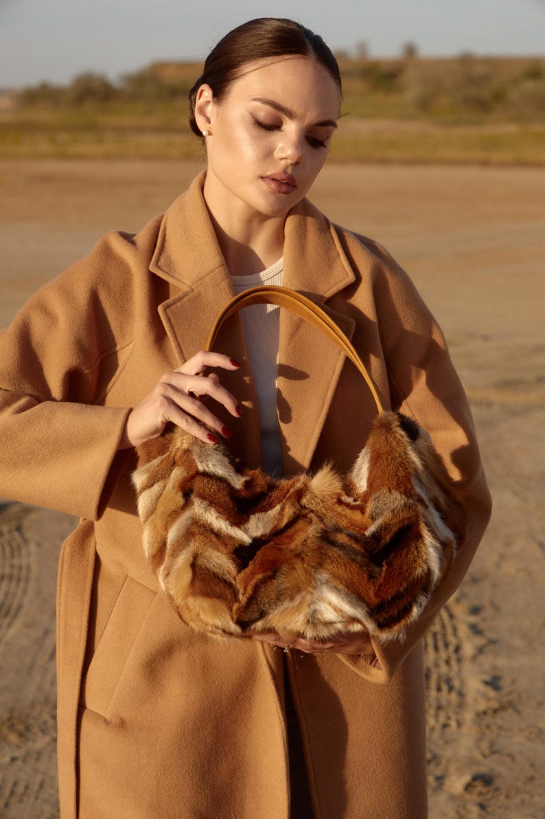 May include: A woman wearing a camel-colored coat holds a brown and white fur handbag with a brown leather strap.