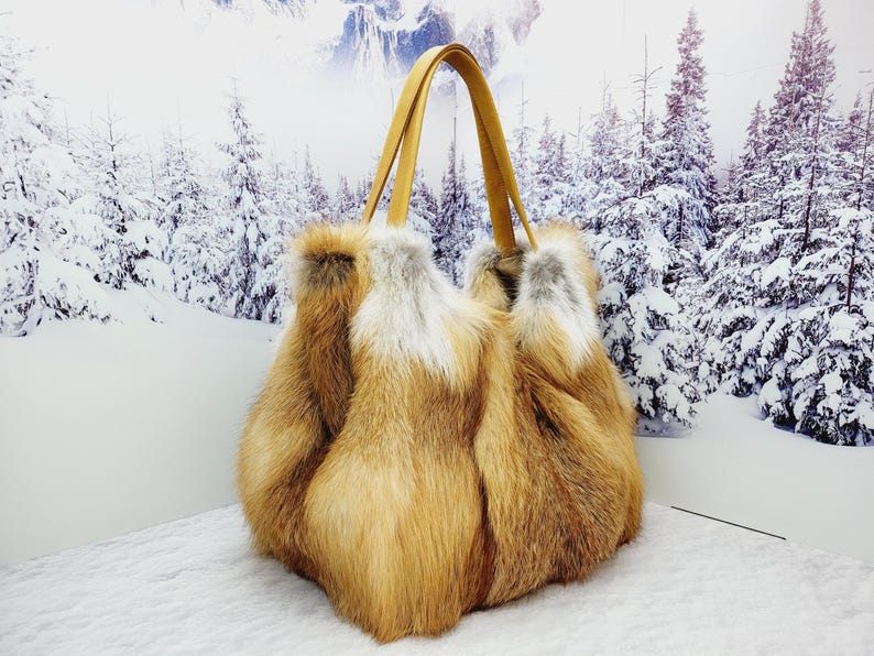 May include: A luxurious tote bag made of reddish-brown and white fur, with tan leather handles. The bag is fluffy and voluminous, perfect for carrying essentials. The background features a snowy winter scene.