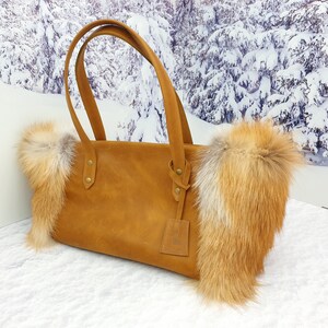 May include: A brown leather tote bag with fox fur trim. The bag has two top handles and a small leather tag hanging from one of the handles.