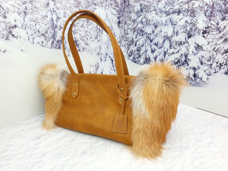 May include: A brown leather tote bag with fox fur trim. The bag has two top handles and a small leather tag hanging from one of the handles.