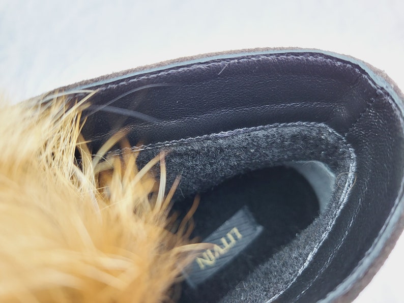 K&ouml;nnte beinhalten: Nahaufnahme des Innenraums eines braunen Wildlederstiefels mit schwarzem Pelzfutter. Der Stiefel hat eine schwarze Ledersohle mit dem in Gold gedruckten Wort "NIALLT".