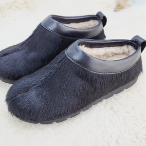 Könnte beinhalten: Ein Paar schwarze, pelzgefütterte Hausschuhe mit schwarzem Lederbesatz. Die Hausschuhe haben ein weiches, flauschiges Futter und eine bequeme Sohle.