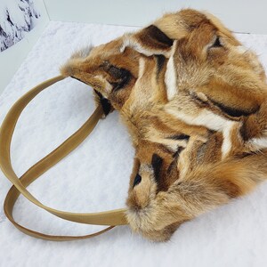 May include: A brown and white fur tote bag with a brown leather strap. The bag is made of fox fur and has a unique pattern.