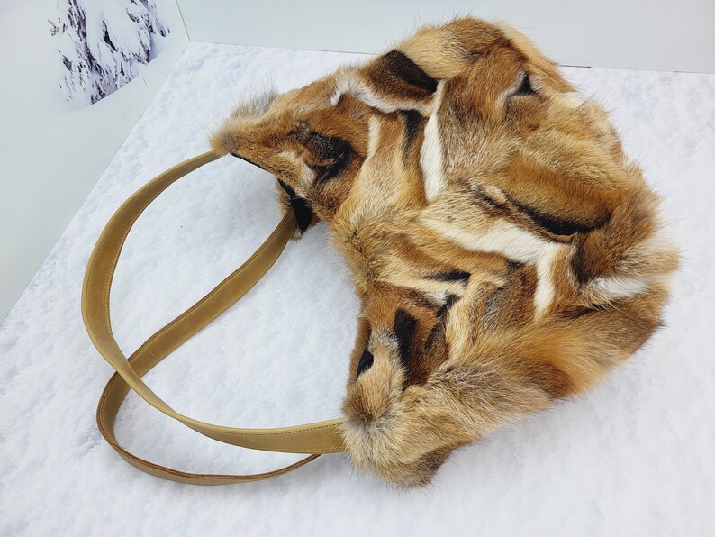May include: A brown and white fur tote bag with a brown leather strap. The bag is made of fox fur and has a unique pattern.