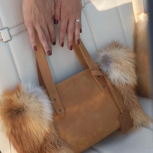 May include: A brown leather tote bag with fox fur trim. The bag has a top handle and a flap closure. The fur is a mix of white and orange.