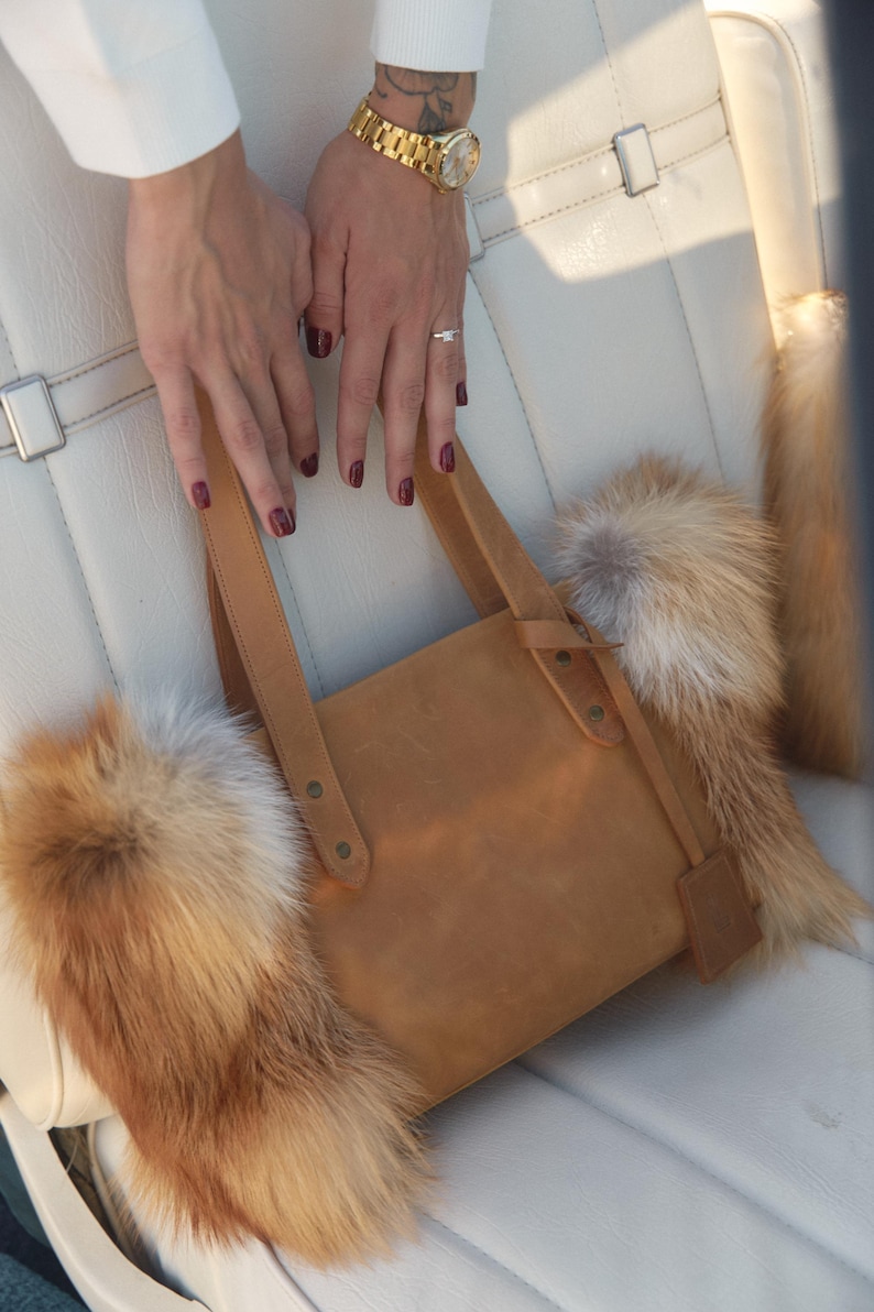 May include: A brown leather tote bag with fox fur trim. The bag has a top handle and a flap closure. The fur is a mix of white and orange.