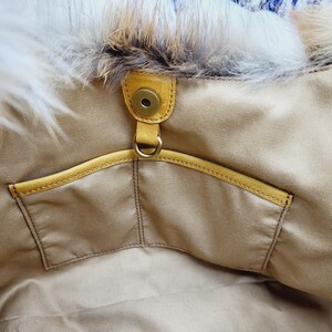 May include: Close-up of a beige handbag interior with a yellow leather pocket and trim. The bag features a magnetic snap closure and a fluffy, white and brown fur trim along the top edge. The bag's interior is a light beige fabric.