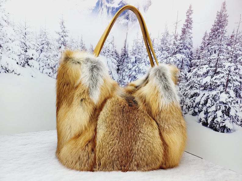 May include: A large, rectangular handbag made of brown and tan fur with a light brown leather handle. The bag is set against a backdrop of a snowy forest scene.