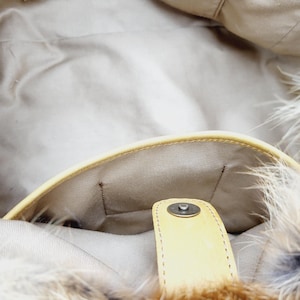 May include: Interior view of a beige handbag with a light yellow leather trim and a magnetic snap closure. The bag's interior is a light beige fabric. The exterior features a fluffy, light brown and white feather trim.