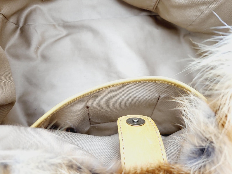 May include: Interior view of a beige handbag with a light yellow leather trim and a magnetic snap closure. The bag's interior is a light beige fabric. The exterior features a fluffy, light brown and white feather trim.
