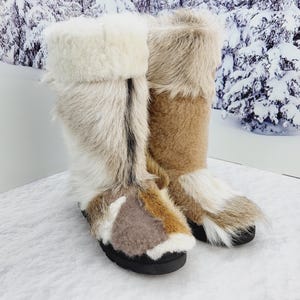 Puede incluir: Un par de botas altas de invierno con exterior de piel. Las botas tienen suela negra y una mezcla de piel beige, marrón y blanca. La parte superior de las botas está doblada, revelando un material cremoso y esponjoso.