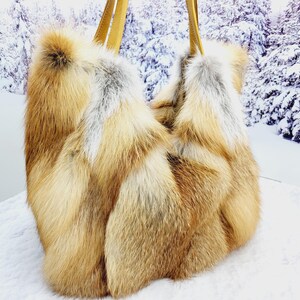 May include: A luxurious, oversized tote bag crafted from natural fox fur. The bag features a mix of golden brown, cream, and gray fur, with two tan leather handles. The bag's design is a soft, unstructured shape, perfect for carrying essentials.