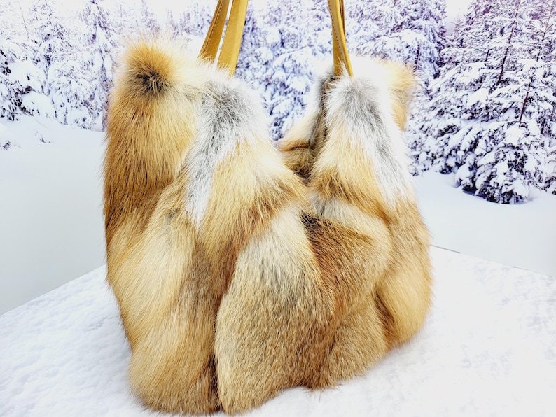 May include: A luxurious, oversized tote bag crafted from natural fox fur. The bag features a mix of golden brown, cream, and gray fur, with two tan leather handles. The bag's design is a soft, unstructured shape, perfect for carrying essentials.