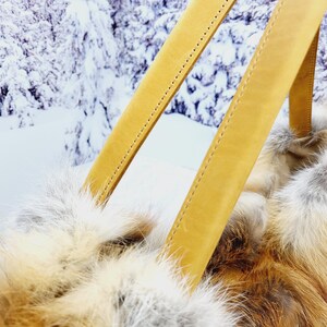 May include: Close-up of a handbag with tan leather straps and a fluffy, fur-like body. The bag's texture is a mix of white, tan, and reddish-brown. The background shows a snowy, winter landscape.
