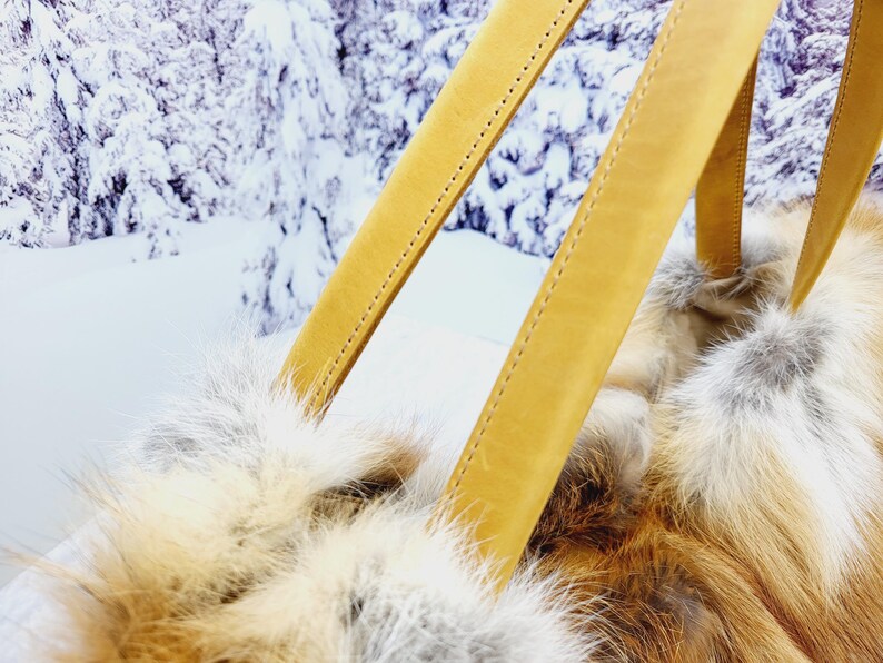 May include: Close-up of a handbag with tan leather straps and a fluffy, fur-like body. The bag's texture is a mix of white, tan, and reddish-brown. The background shows a snowy, winter landscape.