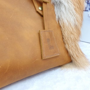 May include: A brown leather tote bag with a fur trim. The bag has a leather tag with the letter "L" embossed on it.