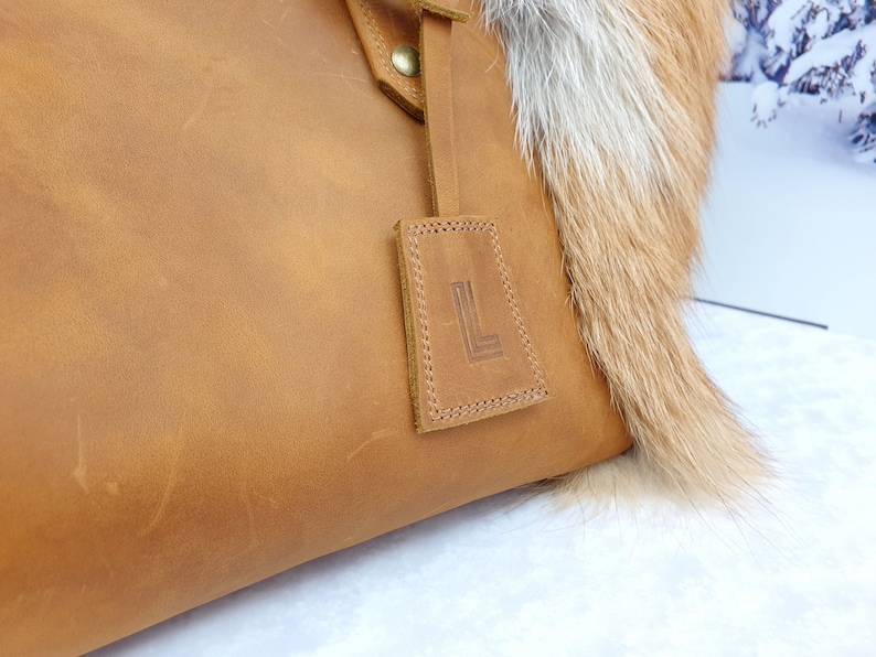 May include: A brown leather tote bag with a fur trim. The bag has a leather tag with the letter "L" embossed on it.