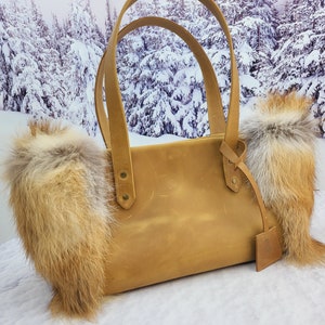 May include: A tan leather tote bag with two fox fur trim accents. The bag has a top handle and a small leather tag.