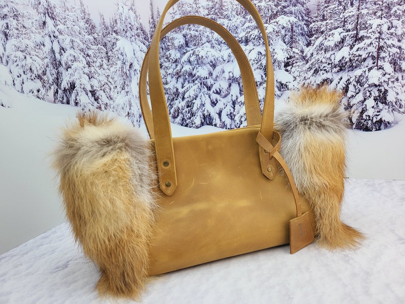 May include: A tan leather tote bag with two fox fur trim accents. The bag has a top handle and a small leather tag.