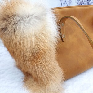 May include: A brown leather tote bag with a fox fur trim. The bag has a long strap and a top zipper closure.